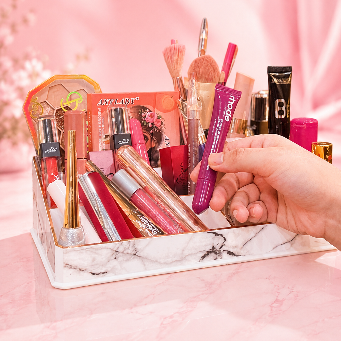 A hand holds a purple tube of ECLAT BY TUBA lip gloss in a white marble cosmetic organizer filled with various makeup items, set against a pink background.
