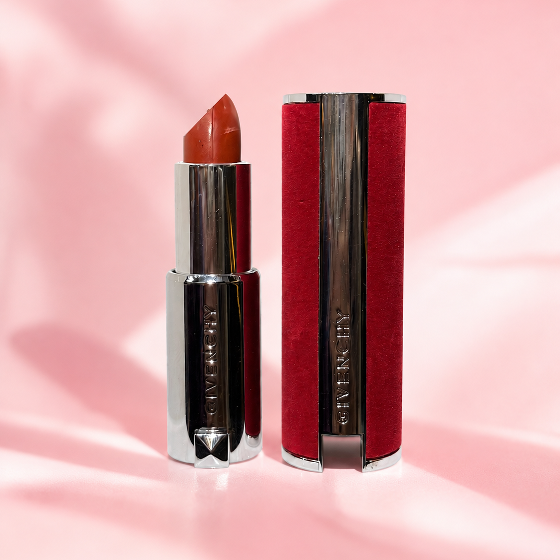 A Givenchy lipstick in a warm terracotta shade, shown open next to its red and silver case on a pink satin background. The Givenchy logo is embossed vertically on the silver casing.
