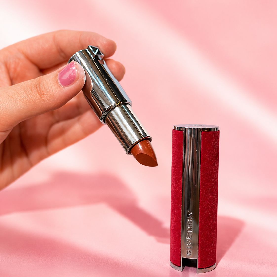 A hand holds an open tube of lipstick showing a warm terracotta-brown shade, next to the closed lipstick case which has a textured red velvet exterior and a silver metallic center column. The background is a soft pink fabric backdrop.