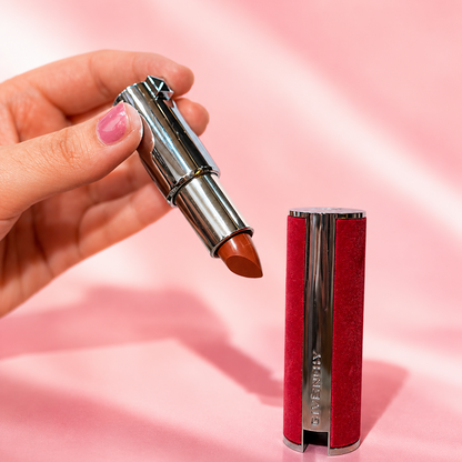 A hand holds an open tube of lipstick showing a warm terracotta-brown shade, next to the closed lipstick case which has a textured red velvet exterior and a silver metallic center column. The background is a soft pink fabric backdrop.