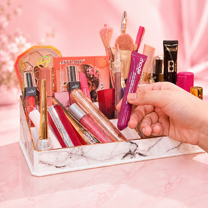 A hand holds a purple tube of ECLAT BY TUBA lip gloss in a white marble cosmetic organizer filled with various makeup items, set against a pink background.